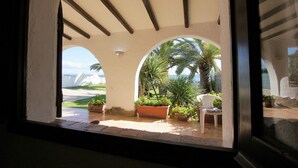 A view through an arched window of a patio area with a white chair, potted plants, and a palm tree. - Villa Mary in Pula: exclusive location with private access to the beach (Pula)