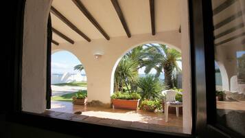 Vista attraverso una finestra ad arco su un patio con una sedia bianca, piante in vaso e una palma.