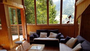 A cozy wooden cabin interior with a sofa, a small table, and a view of a forested valley through large windows.