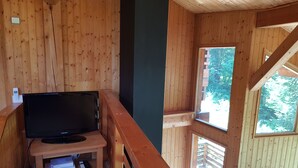 A wooden cabin with a television on a wooden table, a small shelf, and a window overlooking greenery.