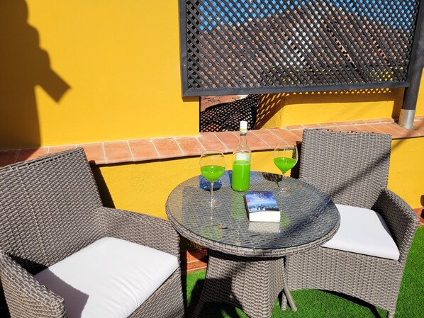 Un pequeño espacio al aire libre con sillas de mimbre y una mesa de vidrio, con una botella y vasos.