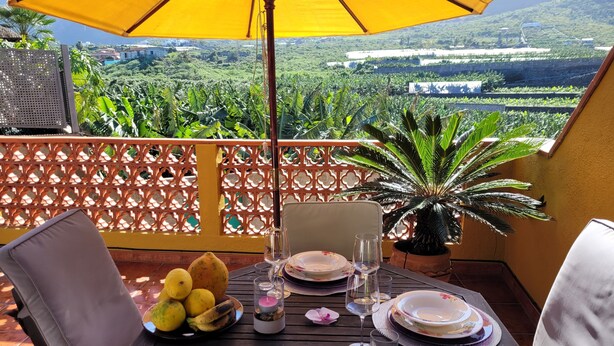 A table set for two with a yellow umbrella, a potted plant, and a scenic view of a lush landscape.