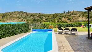 Une piscine rectangulaire à l’eau d’un bleu limpide, entourée d’une terrasse carrelée. Au fond, on aperçoit une colline verdoyante, parsemée d’arbres, et un ciel dégagé.