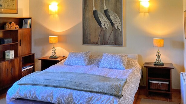 Une chambre avec un plafond en bois, un lit recouvert d’une literie à motifs, deux lampes de chevet et un tableau représentant des oiseaux, accroché au mur.