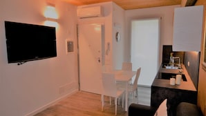 Een moderne woonkamer met een flatscreen televisie, een eethoek met witte stoelen, en een keuken met een fornuis en een spoelbak.