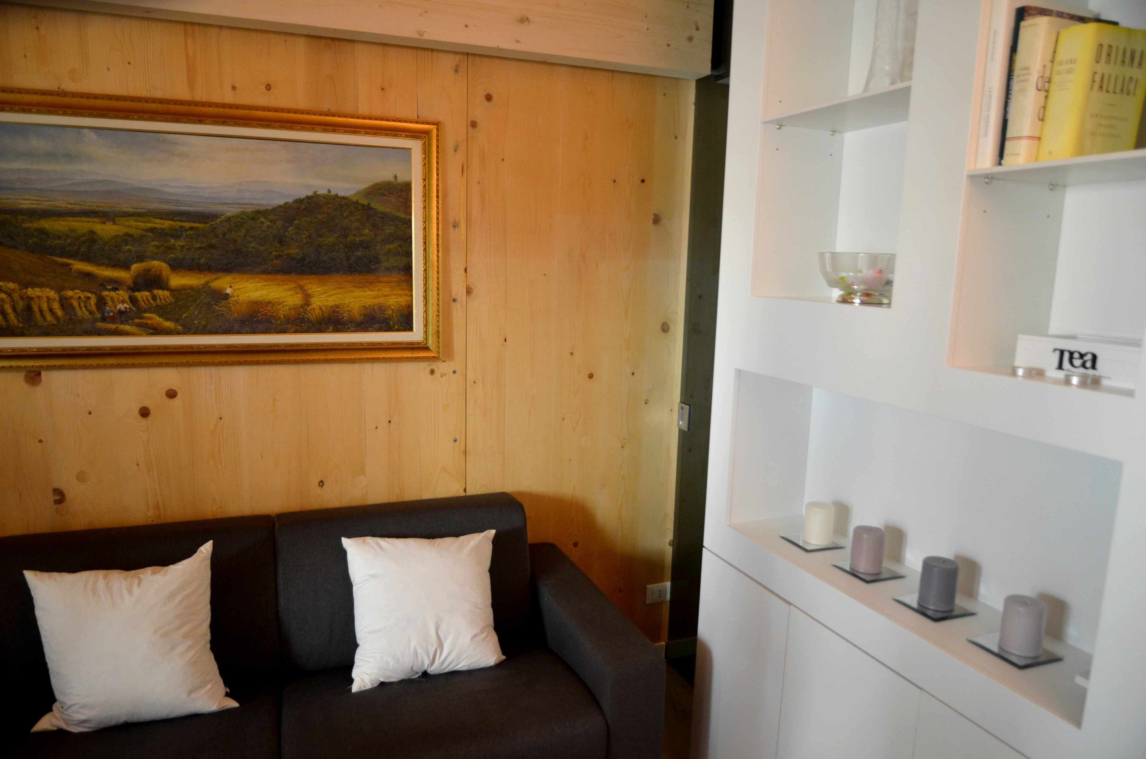 A room with a wooden wall, a painting of a landscape, a dark-colored sofa with white cushions, and a white shelf with tea candles and books.