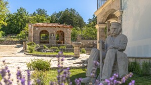 A stone building with a tiled roof, a statue of a seated man, and a stone pathway leading to a small structure with arches. - Villa Marchelina Grubine - Beautiful stone villa with 2 pools (Grubine)