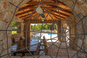 Un espacio de estar al aire libre cubierto, con paredes de piedra y techo de madera, con vistas a una piscina.