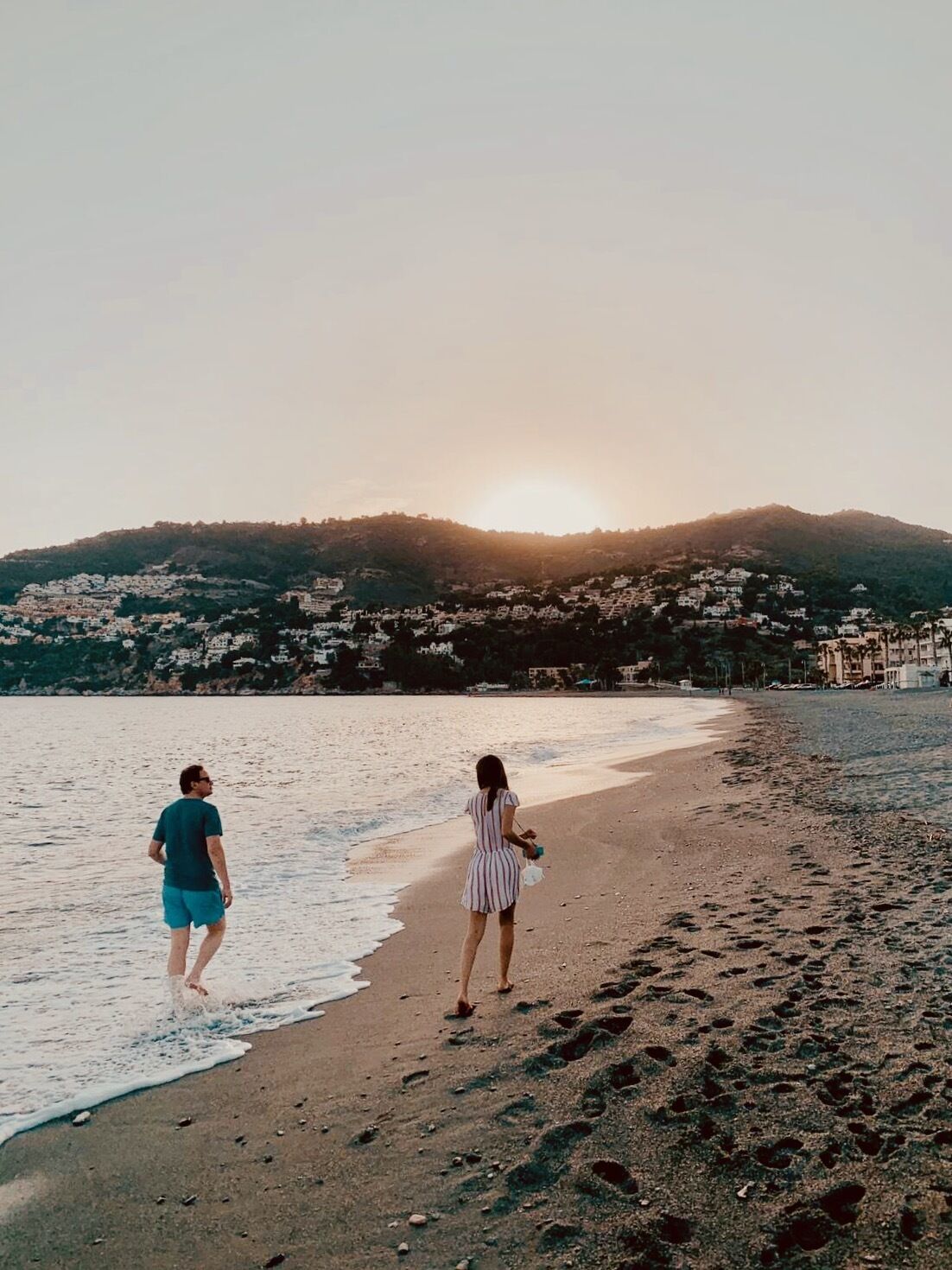 A beach with footprints leading to the water, a sunset in the background, and a hilly landscape with buildings.