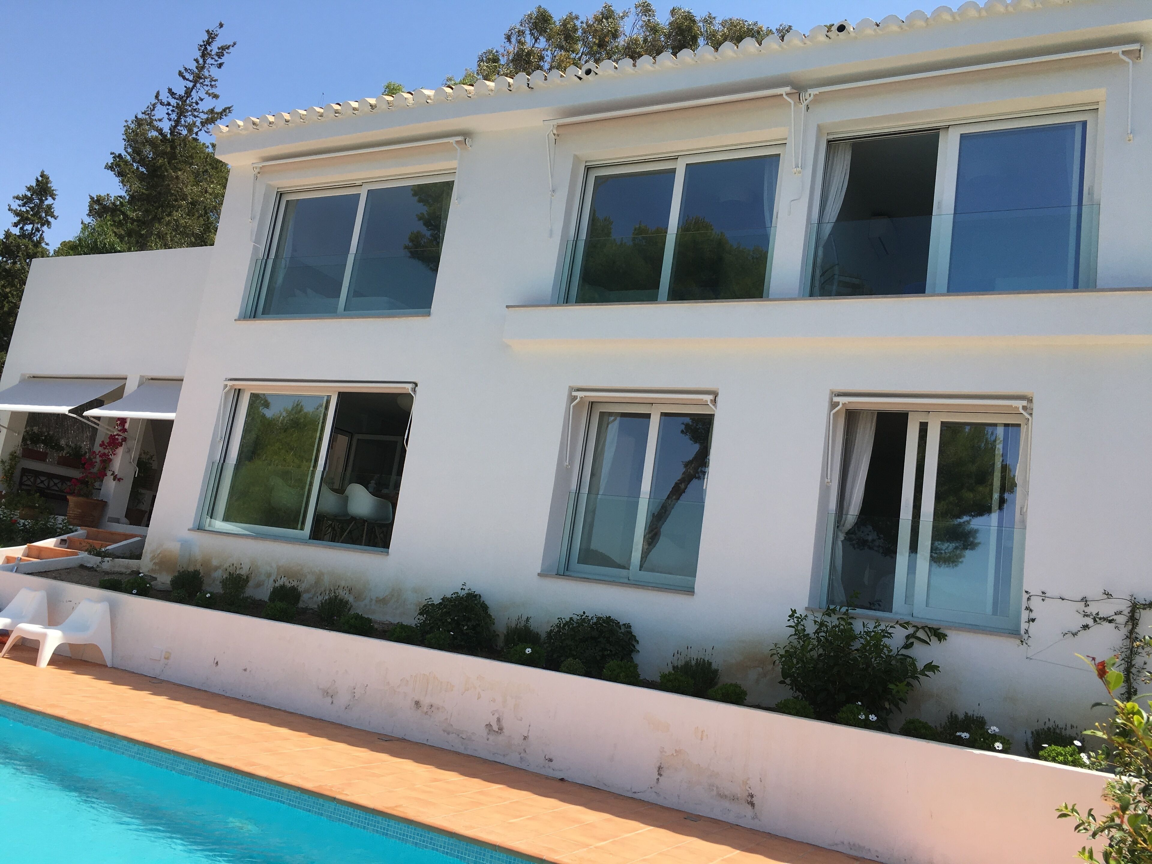 Une maison moderne à deux étages, dotée d’une piscine, de grandes fenêtres et d’un balcon aménagé avec du mobilier d’extérieur.