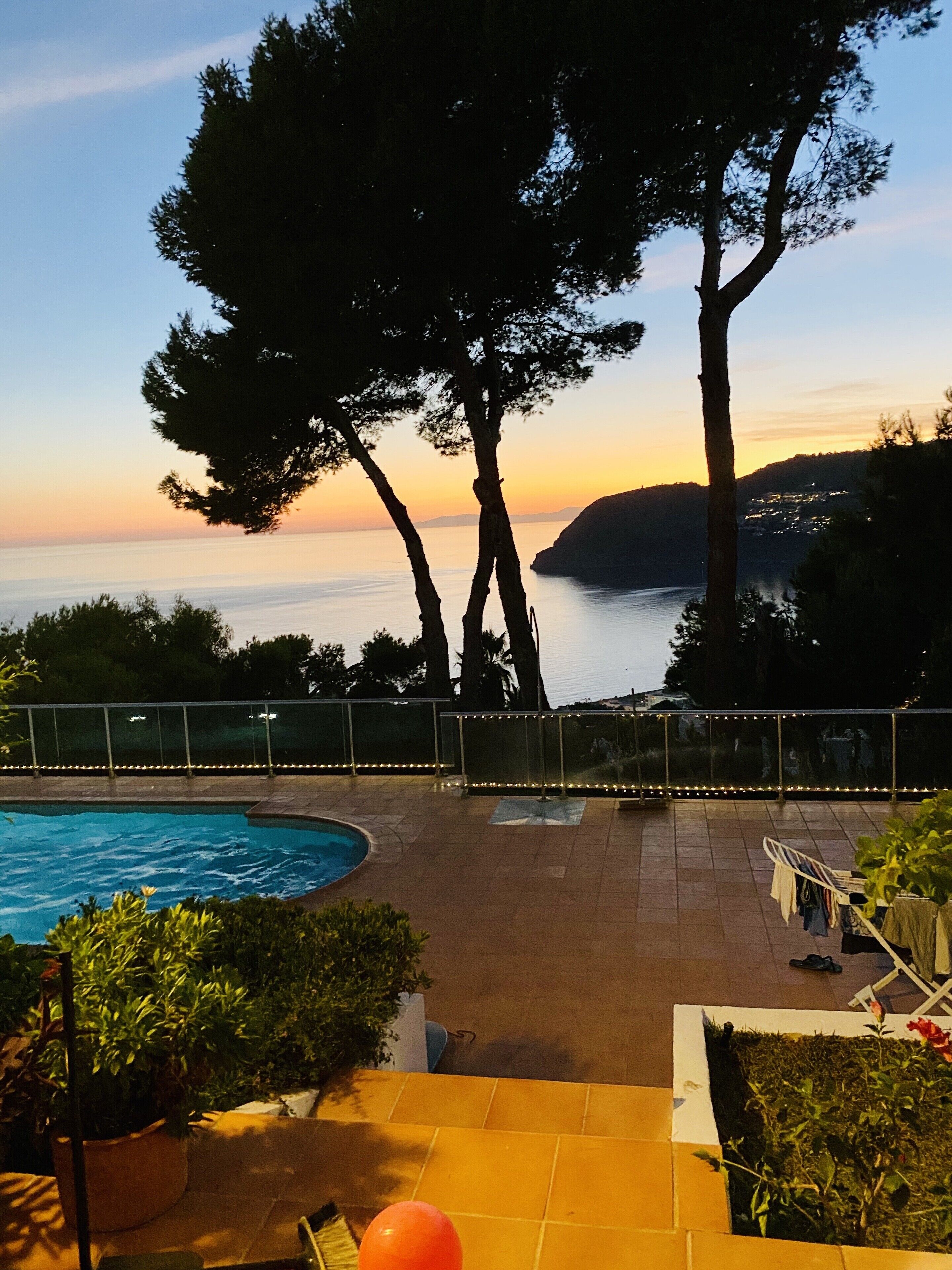 Zona de piscina con vista al mar al atardecer, rodeada de árboles y una barandilla.