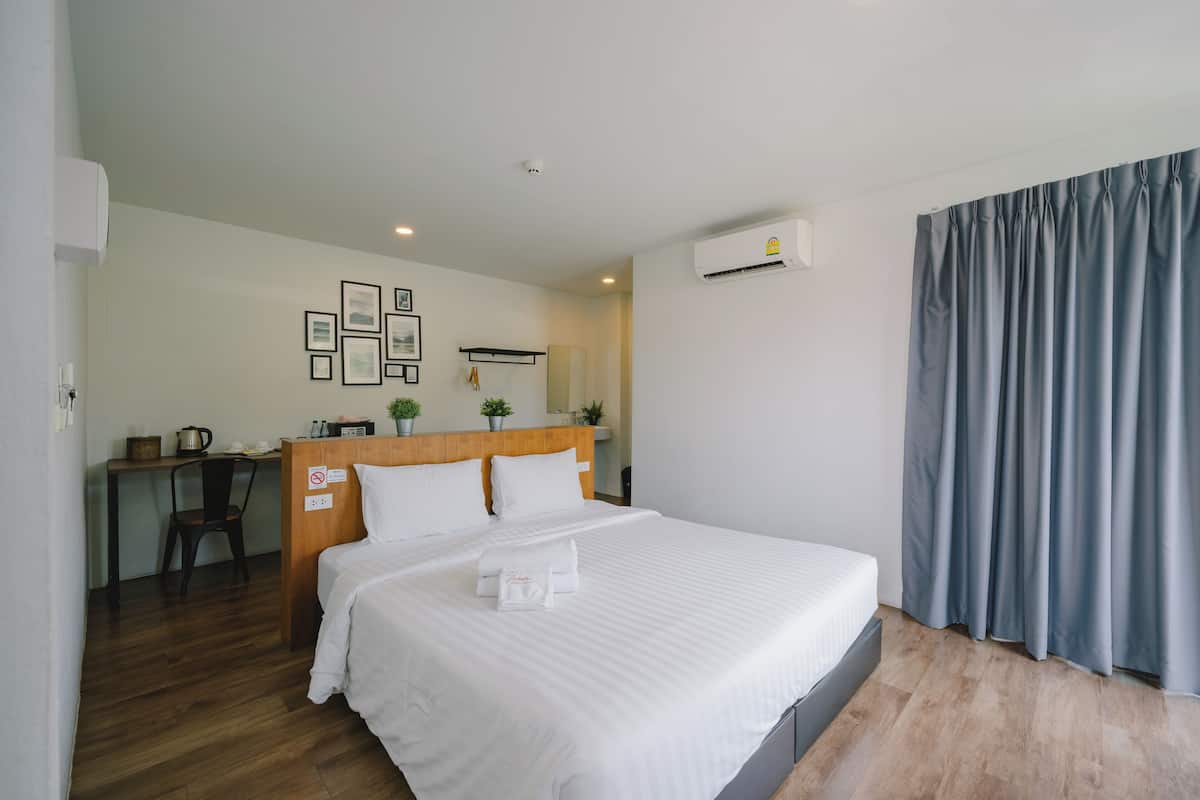 A hotel room with a bed, a desk, and a chair. There is a wall-mounted air conditioner and a window with curtains.