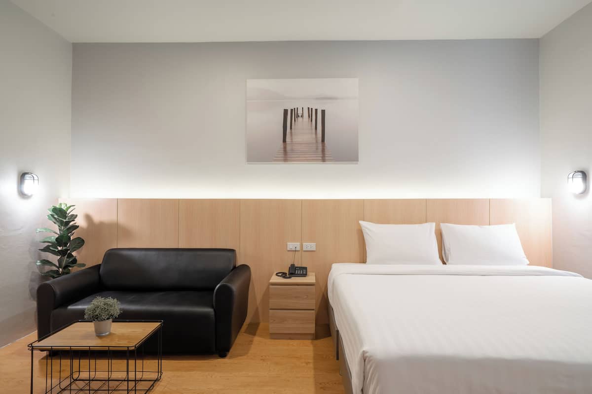 A modern hotel room with a wooden headboard, a black leather sofa, a small wooden table with a plant, a bedside table with a phone, and a large bed with white linens.