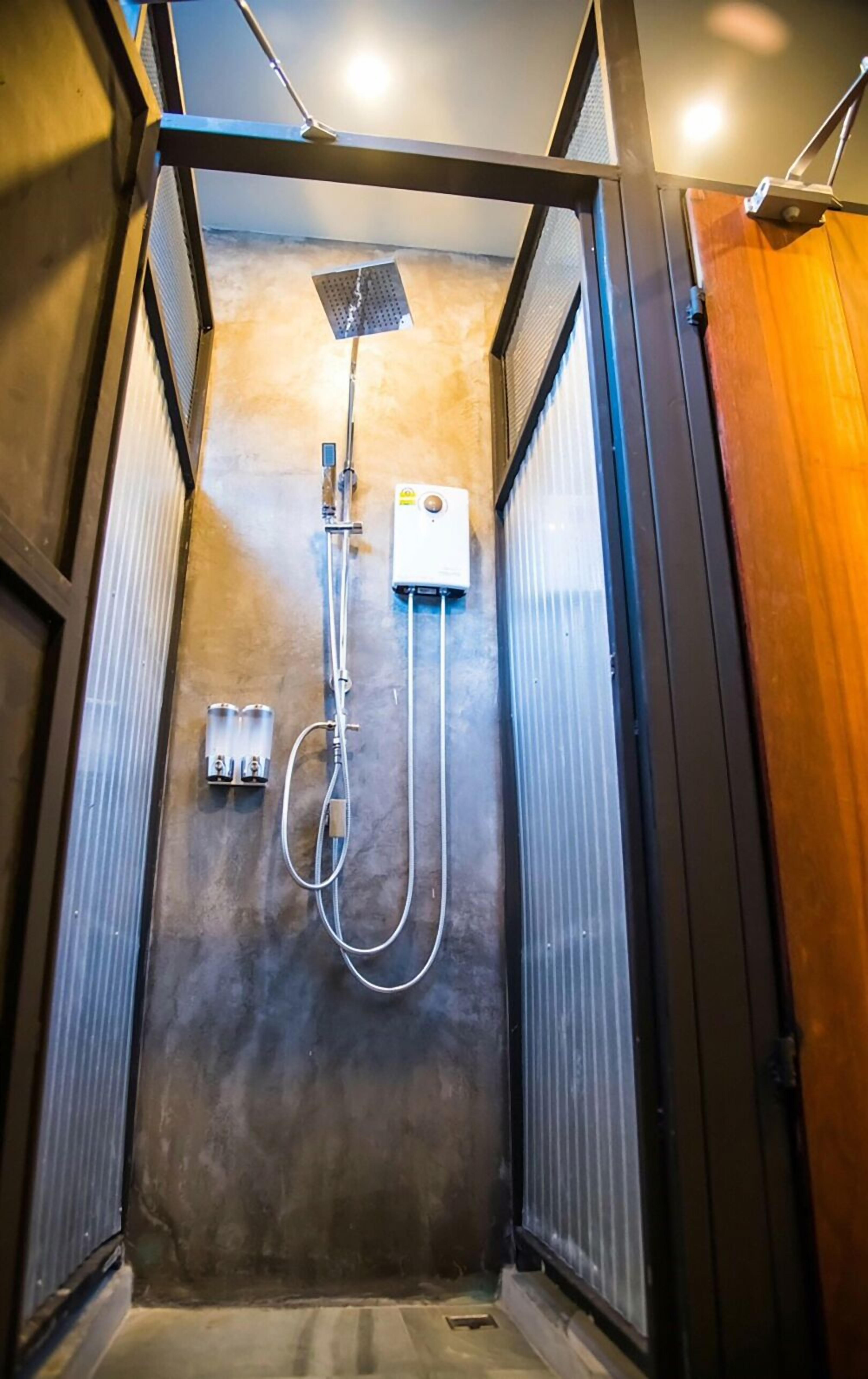 Un box de ducha pequeño y de estilo industrial con puerta metálica, un cabezal de ducha de mano y un panel de control en la pared.