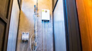 Un box de ducha pequeño y de estilo industrial con puerta metálica, un cabezal de ducha de mano y un panel de control en la pared.