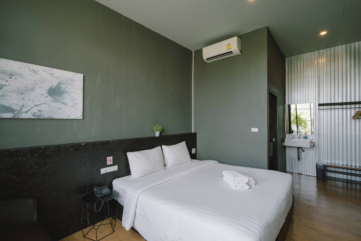 A modern hotel room with a large bed, a bedside phone, a small table with a chair, a wall-mounted air conditioner, and a window with a view of greenery.