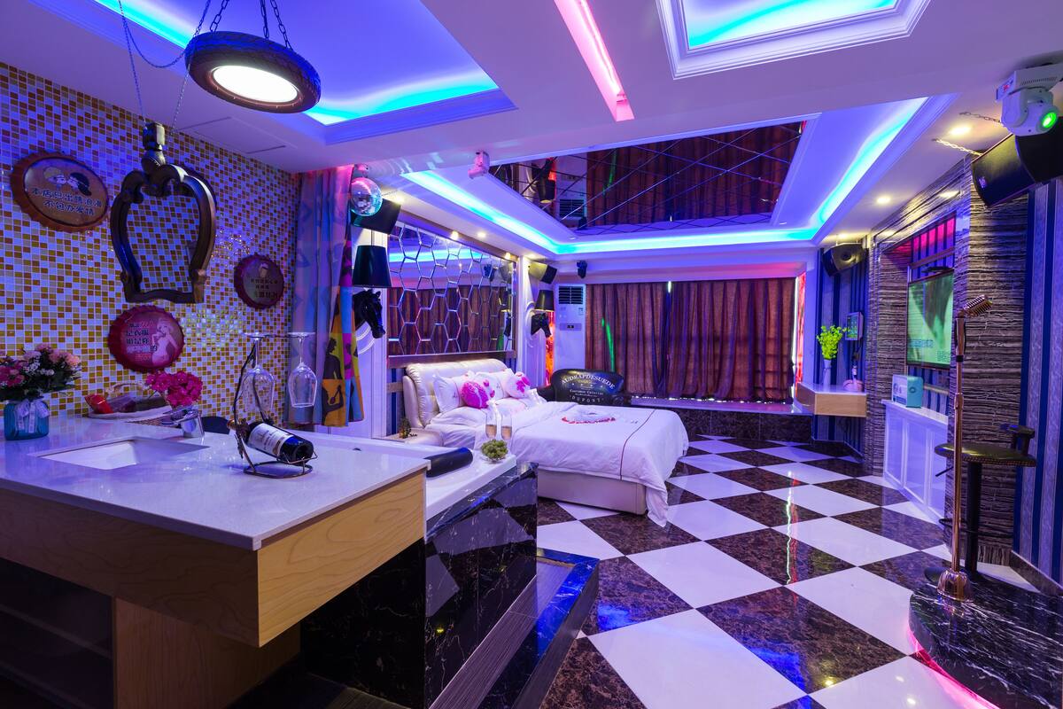 A hotel room with a checkered floor, a bed with white bedding, a vanity with a mirror, and a ceiling with blue and white lighting.