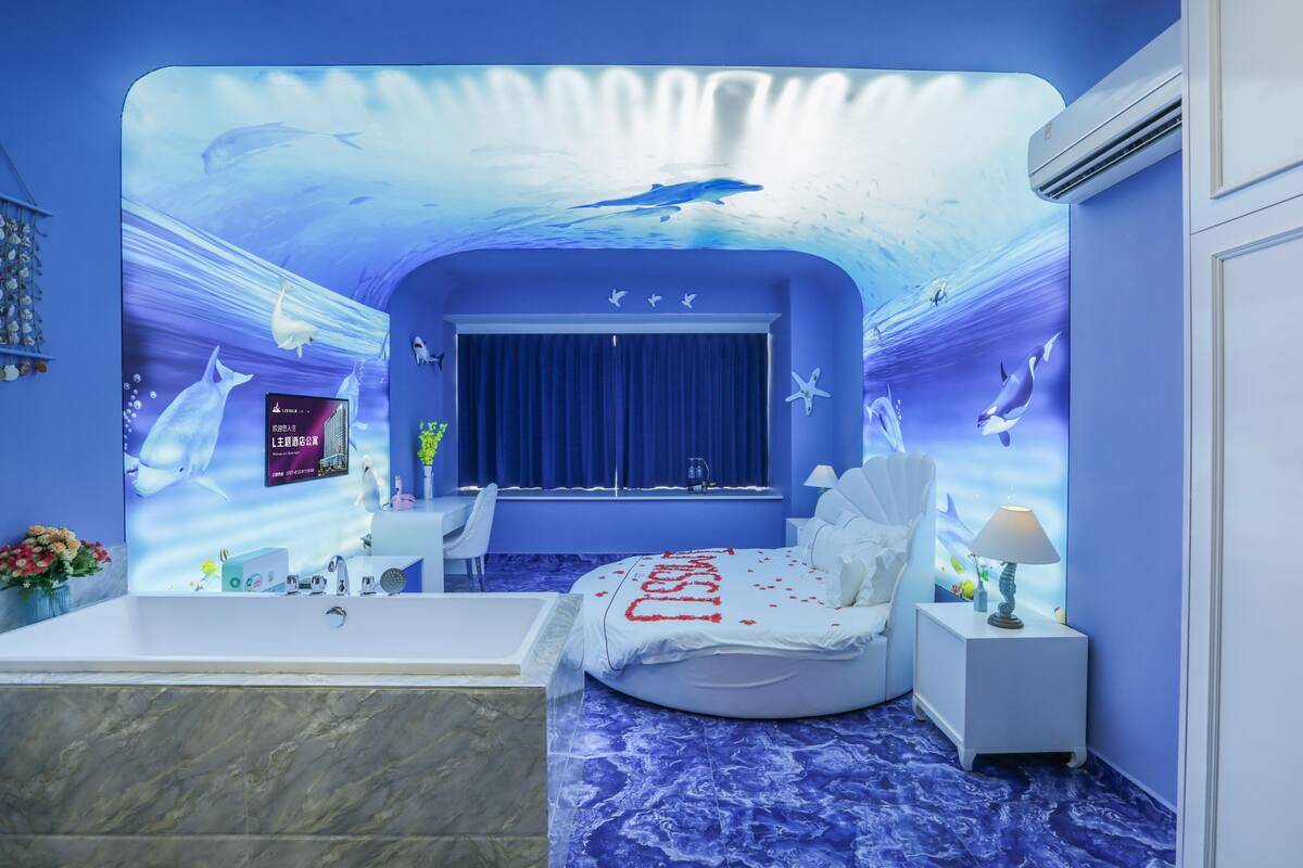 A bedroom with a blue ocean-themed mural, a bed with a white bedspread, a bathtub, and a nightstand with a lamp.