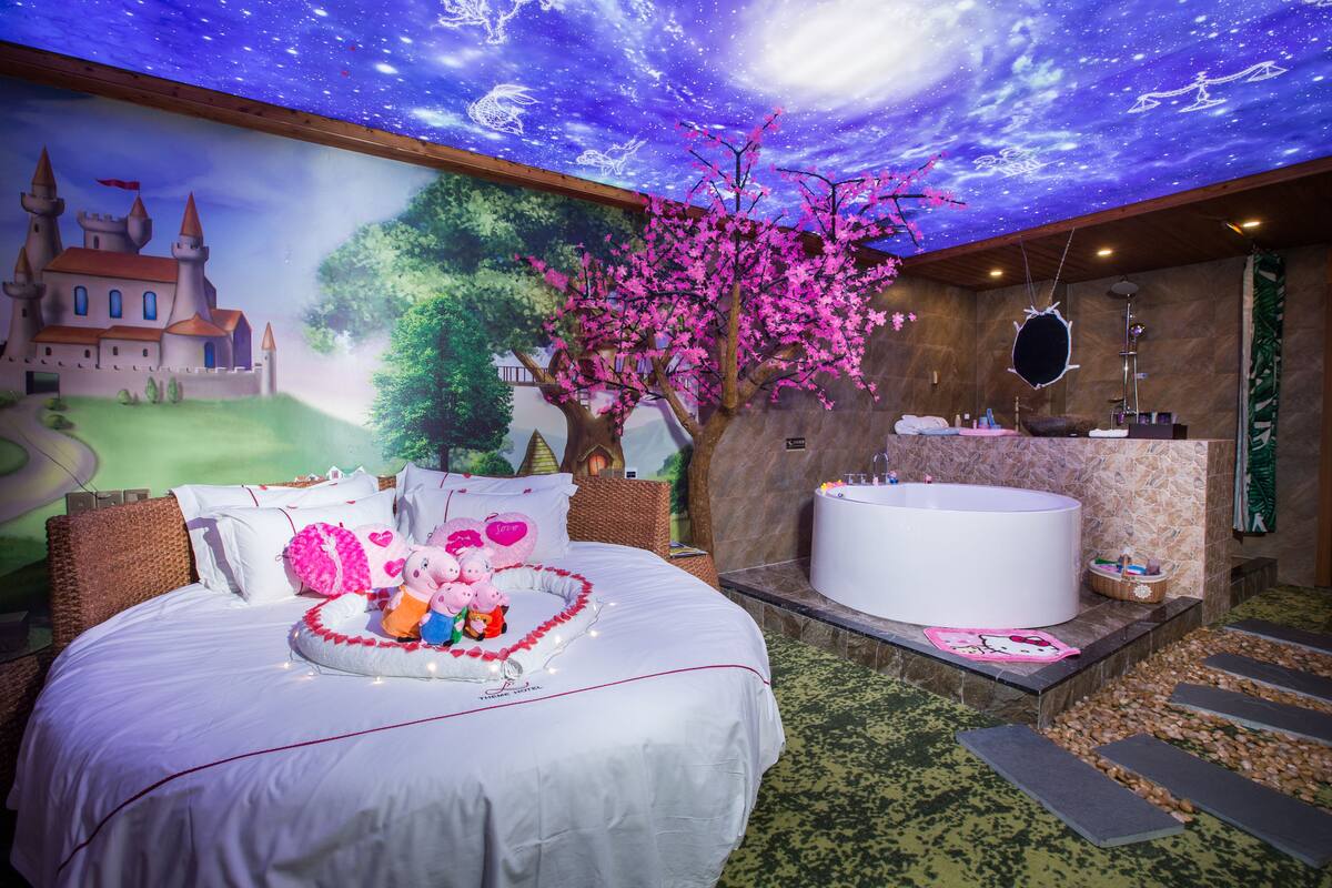 A bedroom with a large bed, a night sky mural, a tree, and a bathroom with a bathtub and sink.