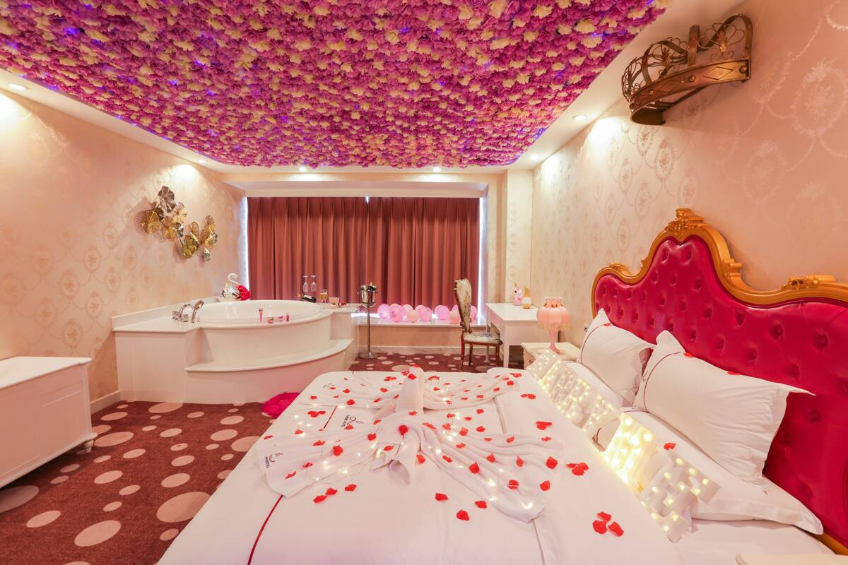 A luxurious bedroom with a bed adorned with rose petals and lights, a large mirror, and a pink headboard.