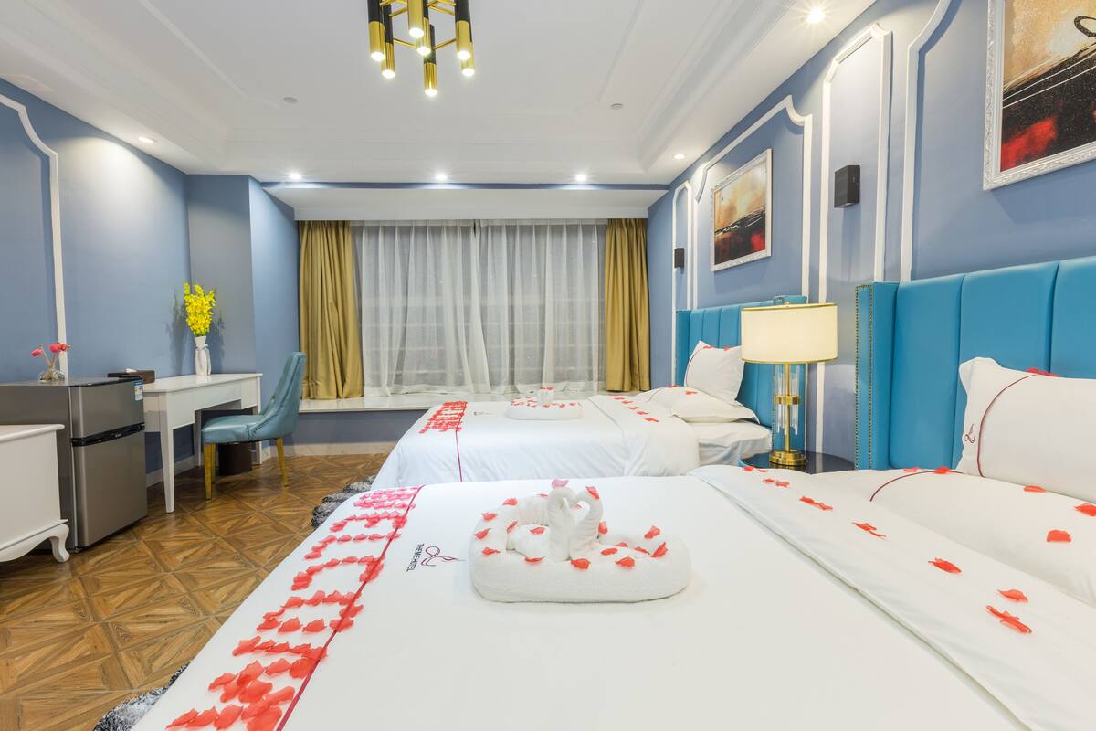 A hotel room with two beds, a desk, a chair, a minibar, and a window with curtains.