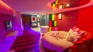 Une chambre d’hôtel moderne avec un plafond incurvé, un grand lit parsemé de pétales de rose, un canapé rouge et une petite table avec des boissons.