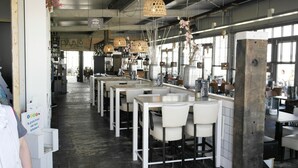 A modern restaurant with white tables and chairs, hanging wicker lamps, and a view of the city through large windows.