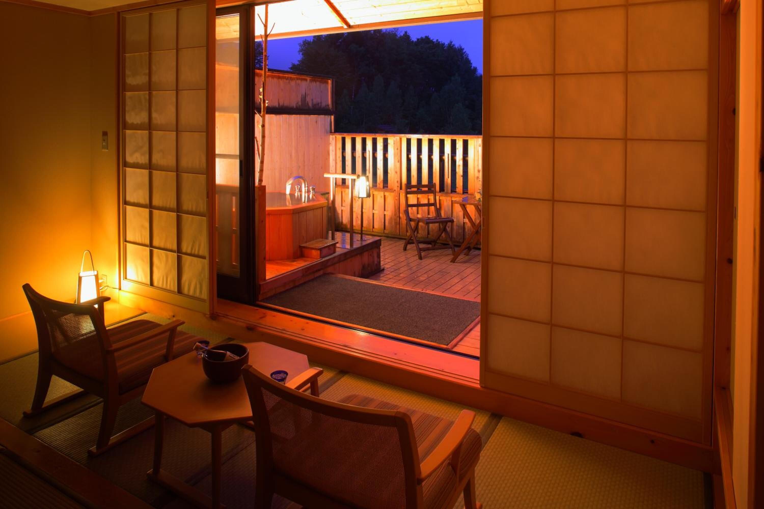 A traditional Japanese room with tatami flooring, a low table, and a sliding door leading to an outdoor deck with a wooden fence and a hot tub.