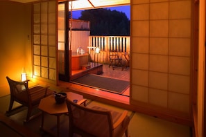 Et traditionelt japansk rum med tatamimåtter, et lavt bord og en skydedør, der fører til en udendørs terrasse med et træhegn og et boblebad.