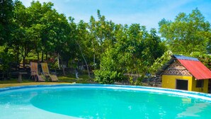 Una piscina con cubierta de madera y sillas de descanso, rodeada de abundante vegetación y un pequeño edificio con techo rojo.