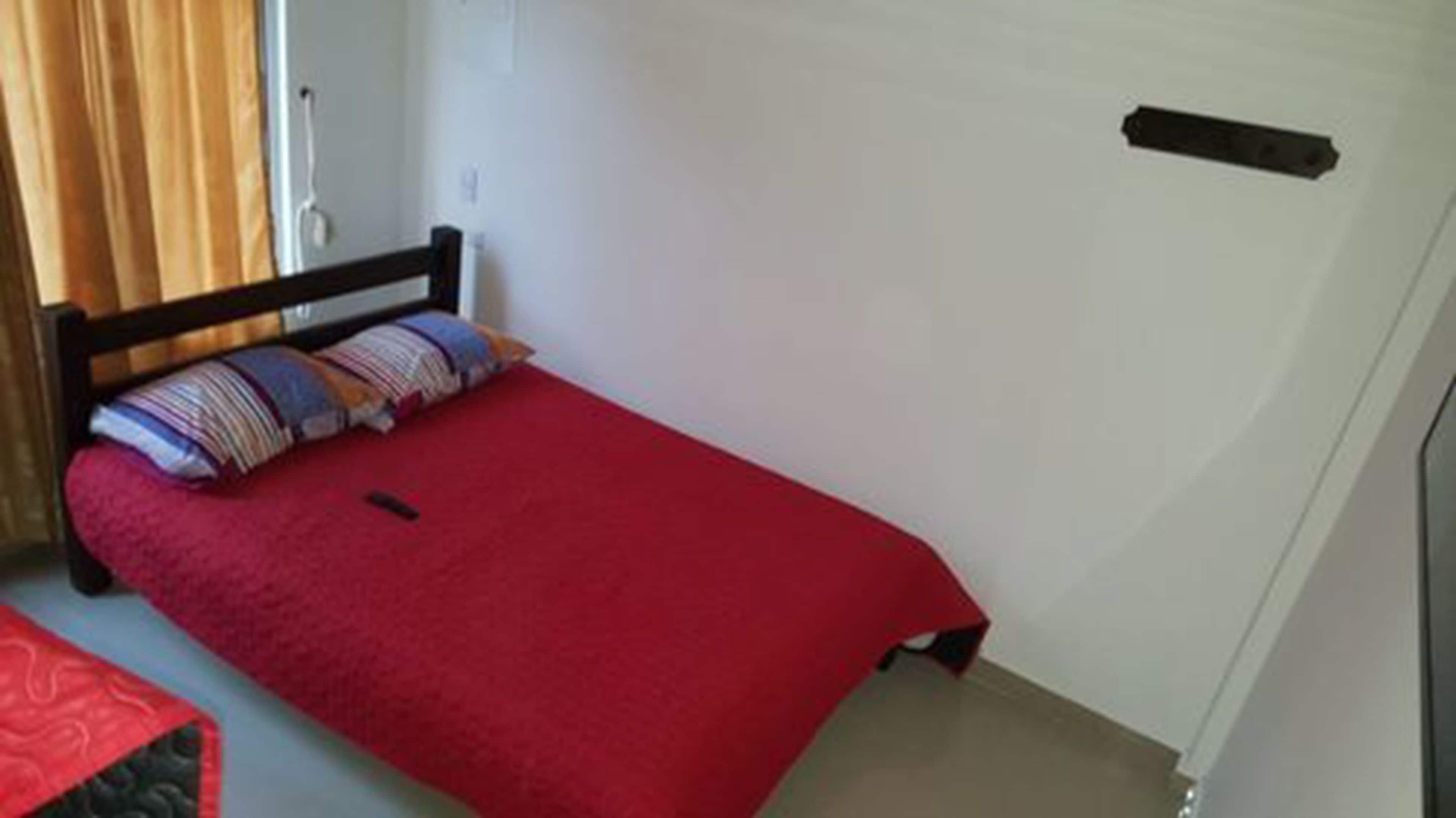 A small, single-bed room with a red bedspread, a wooden headboard, and a window with curtains.