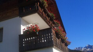 Een gebouw met twee verdiepingen, een witte gevel, houten balkons met rode bloemen en een berg op de achtergrond.