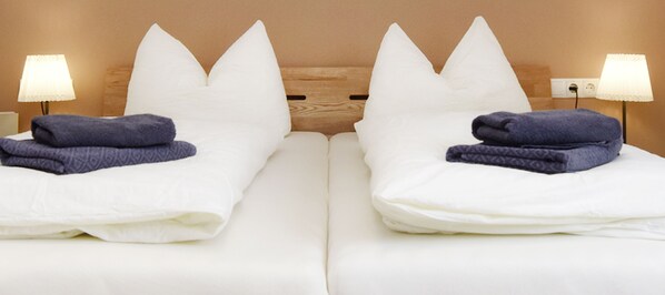 Habitación de hotel con dos camas, cada una con almohadas blancas y toallas azules dobladas. Hay lámparas de noche a ambos lados de las camas.