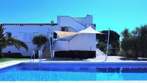 A swimming pool with a white building in the background, a covered patio area, and a staircase leading to the building.