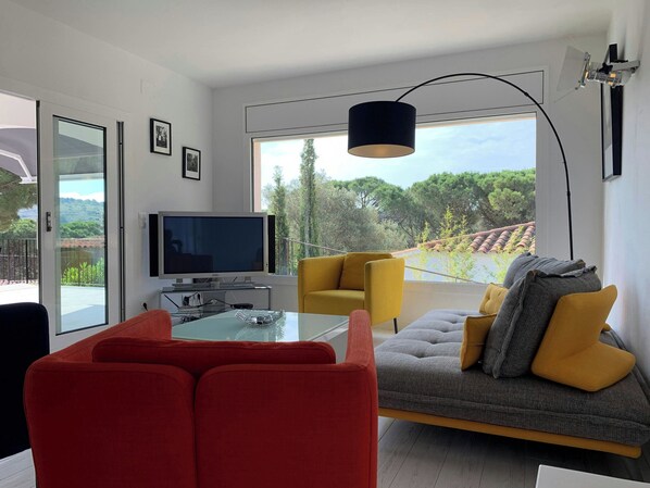 A modern living room with a sofa, chairs, a TV, and a large window with a view of trees.