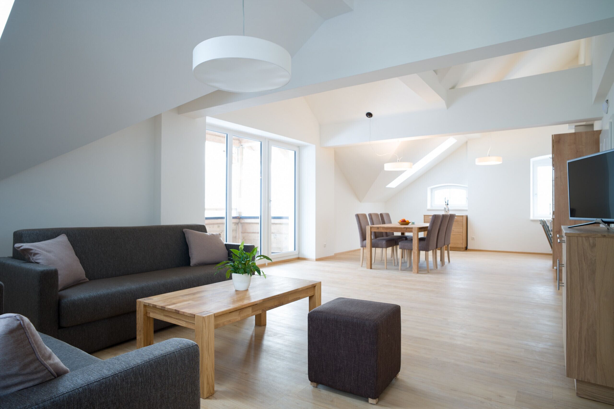 Un salon moderne comprenant un canapé, un pouf et une table basse, un coin repas avec des chaises et une table, et un coin cuisine avec des placards et un téléviseur.
