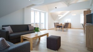 Uma sala de estar moderna com sofá, banqueta e mesa de centro, uma área de refeições com cadeiras e mesa, e uma cozinha com armários e uma televisão.