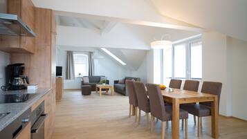 Een moderne keuken met houten kastjes en een inbouwoven. Er is een zithoek met een houten tafel en stoelen. De kamer heeft grote ramen en een schuine kap.