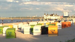 Uma praia com fileiras de cabanas de praia numeradas, uma torre de salva-vidas e um píer ao fundo.