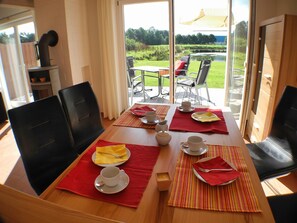 A dining table set with plates, cups, and cutlery, with a view of a garden through large windows.