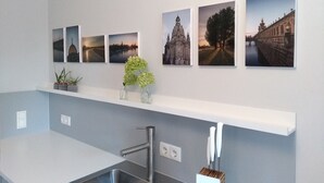 Een keuken met een roestvrijstalen spoelbak, een wit aanrechtblad en een wit schap erboven met vier ingekaderde foto's.