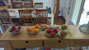 A wooden table with various fruits and vegetables, a bottle, and a vacuum cleaner in a dining area with wooden chairs and a window.