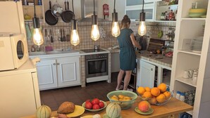 A kitchen with a wooden table displaying fruits and vegetables, a woman cooking, and various kitchen appliances.