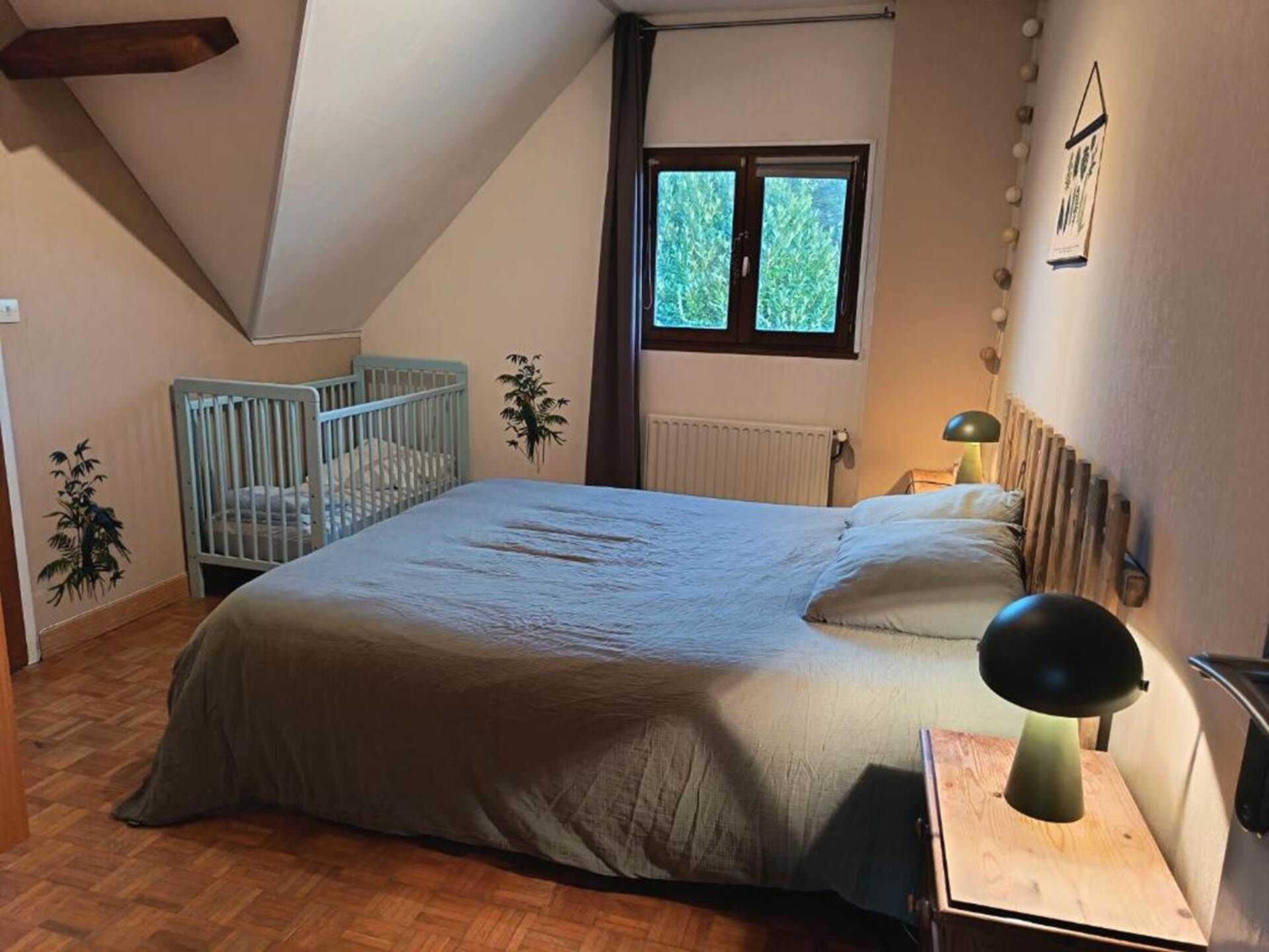 A bedroom with a bed, a crib, a window, a radiator, and a wall-mounted lamp.