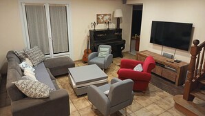 Ein Wohnzimmer mit einer Couch, Sesseln, einem Couchtisch, einem Fernseher, einem Klavier und einer Gitarre.