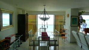 A dining area with a glass table and chairs, a chandelier, and a view of the sea through large windows.
