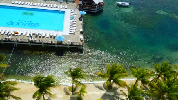A poolside area with a 'Sunrise' sign, a wooden deck, and a beach with palm trees.