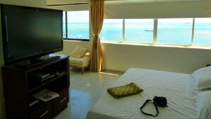 A hotel room with a bed, a TV on a stand, a chair, and a window with a view of the sea.