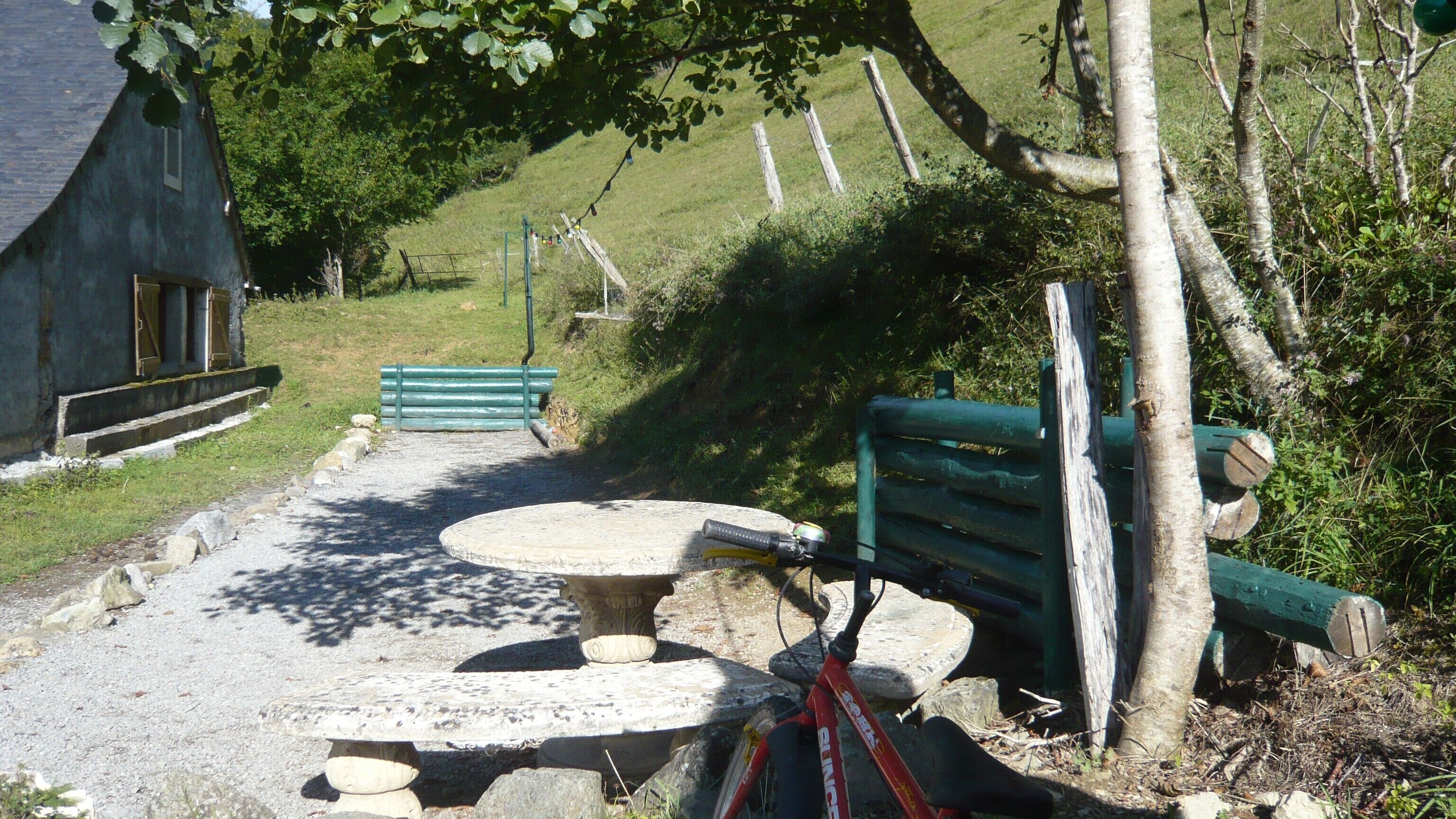 A rustic outdoor seating area with a stone table and benches, surrounded by a gravel path and greenery.