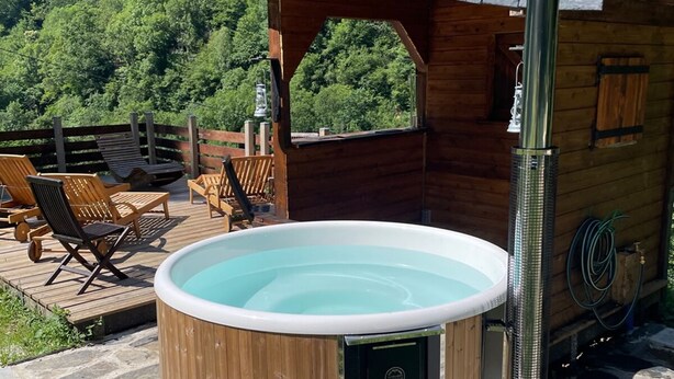 A hot tub with a wooden exterior, a stone deck, and a wooden deck chair area. - Gîte Jaune - House on 5 ha property, swimming pool, sauna, animals, (Osse-En-Aspe)